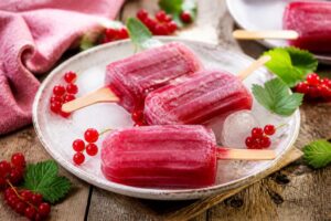 Johannisbeer-Limonaden-Eis am Stiel auf einem Holzbrettchen mit frischen roten Johannisbeeren und einem Glas Limonade im Hintergrund, sommerliches Tageslicht