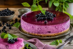 Schwarze-Johannisbeer-Cheesecake ohne Backen auf einem hellen Holztisch – Anschnitt zeigt die violette Fruchtcreme auf dem goldenen Keksboden